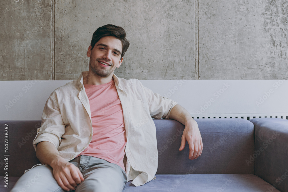 Young fun smiling happy cheerful caucasian man 20s wear casual clothes beige shirt pink t-shirt look camera sitting on grey sofa resting indoors at home on weekends. People lifestyle leisure concept Young fun smiling happy cheerful caucasian man 20s wear casual clothes beige shirt pink t-shirt look camera sitting on grey sofa resting indoors at home on weekends. People lifestyle leisure concept