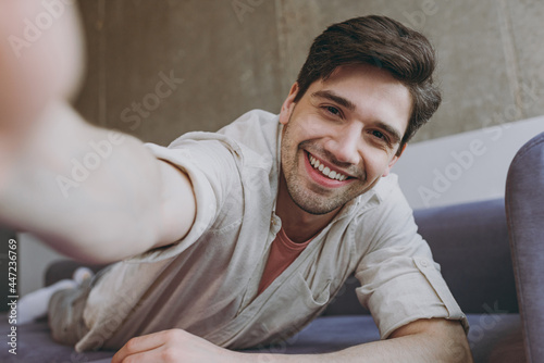 Tablou pe pânză Close up young fun cheerful happy man 20s wearing casual clothes beige shirt pink t-shirt doing selfie shot on mobile phone lying on grey sofa rest indoors at home on weekends