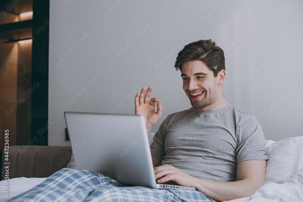 Young man in pajamas grey t-shirt lying sit in bed talk by video call use work laptop pc computer show okay gesture rest relax at home indoors bedroom. Good mood sleeping night morning bedtime concept Young man in pajamas grey t-shirt lying sit in bed talk by video call use work laptop pc computer show okay gesture rest relax at home indoors bedroom. Good mood sleeping night morning bedtime concept