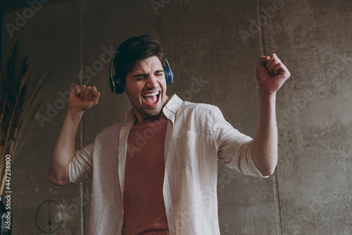 Young overjoyed satisfied student happy european positive man wear casual clothes beige shirt pink t-shirt headphones dancing listen music rest indoors at home on weekends on brown wall background