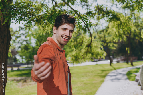 Side view young man in orange jacket walking stretch hand to camera say come with me call rest relax in spring green city park go down alley sunshine lawn outdoors on nature. Urban leisure concept photo