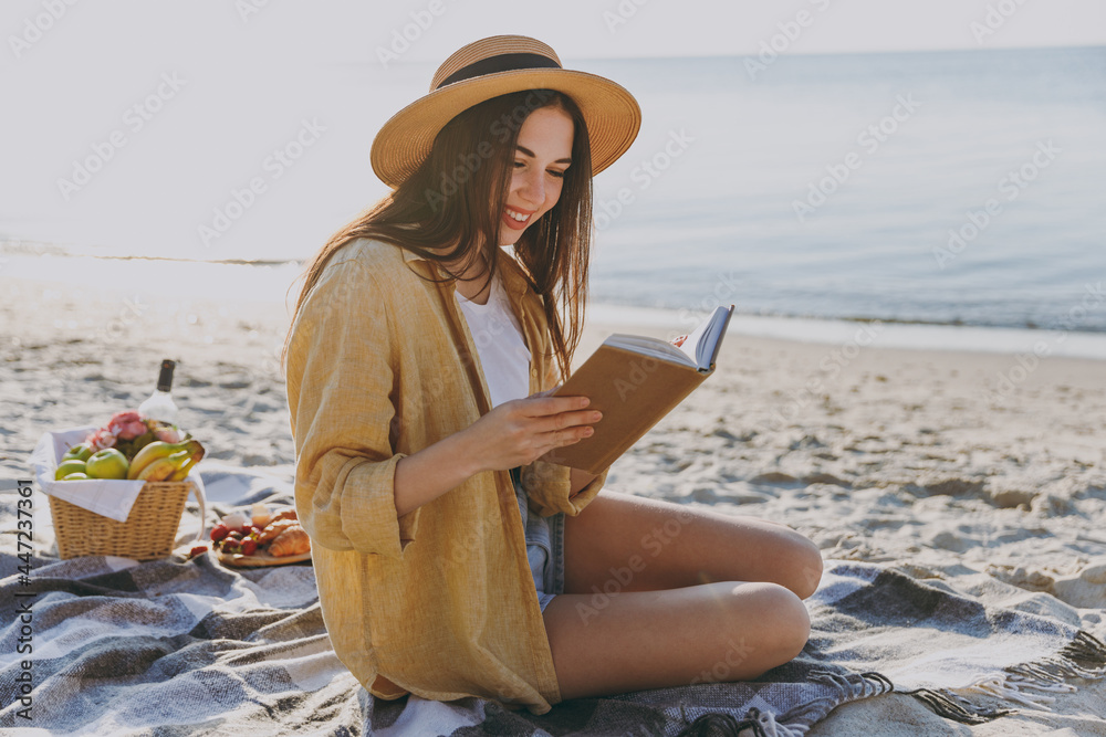 Full size young happy traveler tourist woman in straw hat shirt summer clothes reading book sit on plaid have picnic outdoors on sea sand beach background People vacation lifestyle journey concept