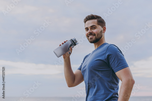 Bottom view young strong sporty athletic fit sportsman man wear sports clothes warm up training drink water hold bottle at sunrise sun dawn over sea beach outdoor on pier seaside in summer day morning