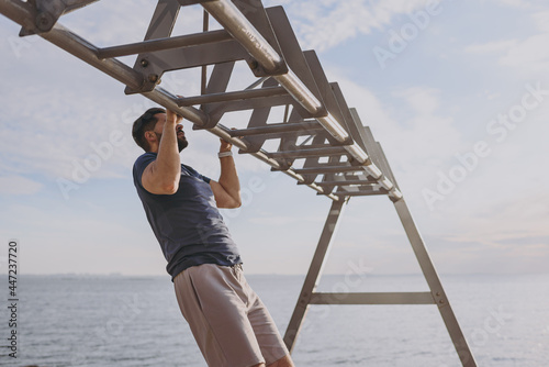 Youngn side view sporty strong toned fit sportsman man wearing sports clothes warm up training pull-up on the horizontal bar at sunrise over sea beach outdoor on pier seaside in summer day morning photo