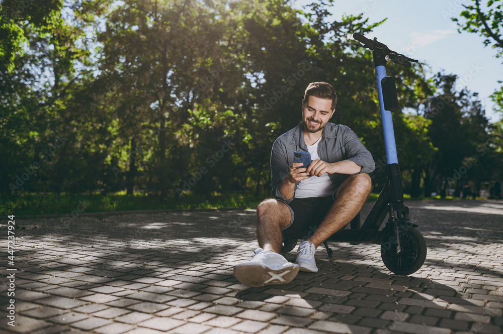 Fototapeta premium Full length bottom view young man in blue shirt sit near riding scooter hold mobile cell phone go down alley rest in spring green city sunshine park outdoors on nature Urban leisure lifestyle concept.