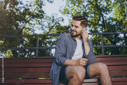 Smiling young man 20s wearing blue shirt shorts earphones sit on bench use mobile cell phone listen to music rest relax in spring green city park outdoor on nature Urban leisure lifestyle concept.