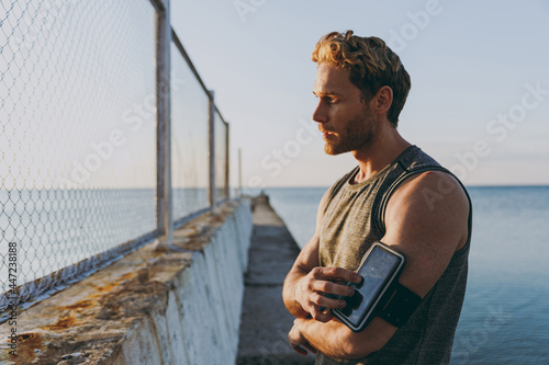 Young side view strong sporty athletic toned fit sportsman man in sports clothes warm up training use mobile cell phone app at sunrise sun over sea beach outdoor on pier seaside in summer day morning © ViDi Studio