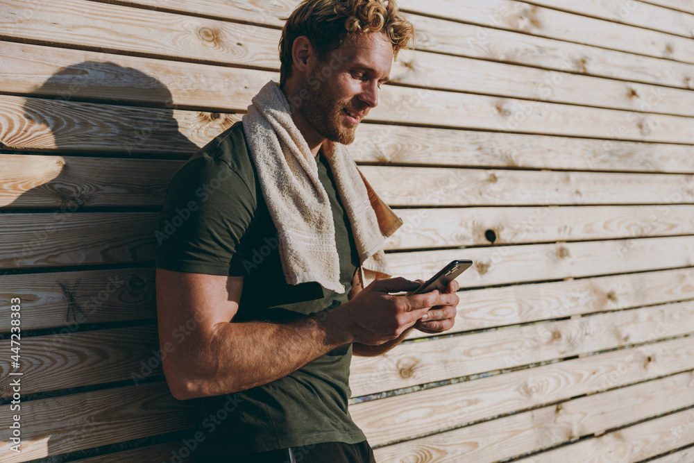Obraz premium Young strong sporty athletic fit sportsman man wear sports clothes towel warm up training use mobile cell phone surfing internet leaning on wooden wall sea beach outdoor seaside in summer day morning.