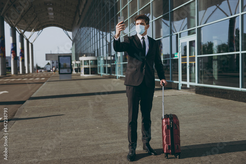 Full body traveler businessman young man in black suit sterile facial mask stand outdoor at international airport terminal with suitcase do selfie shot on mobile cell phone Air flight business concept