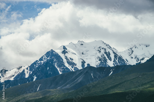 Awesome mountains landscape with high snowy mountain range among white clouds in blue sky. Atmospheric highland scenery with snow-white big mountain ridge in overcast weather. Wonderful snowy pinnacle