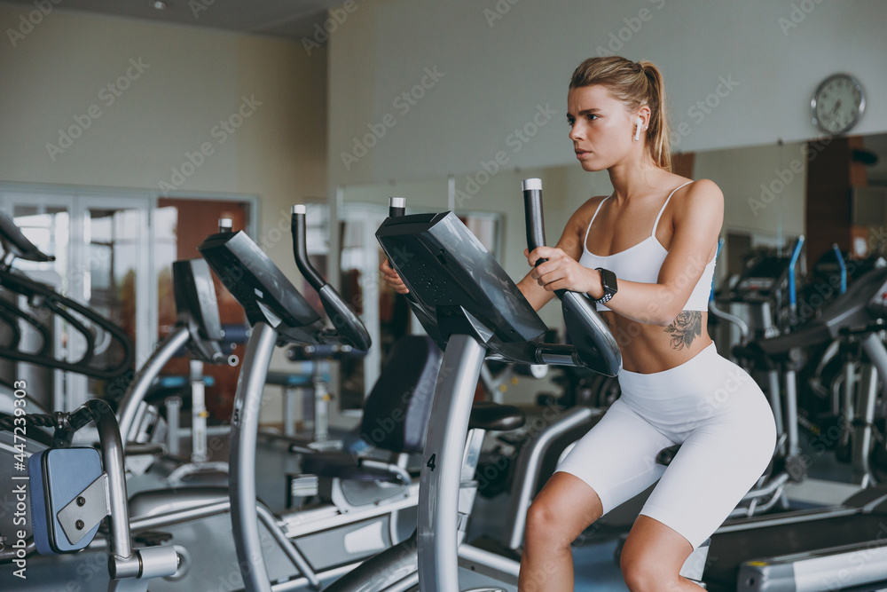Young concentrated serious skinny strong sporty athletic sportswoman caucasian woman in white sportswear earphones listen music warm up train use exercise bike in gym indoors Workout sport concept.