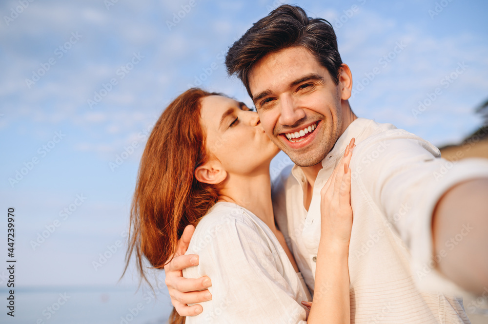Obraz premium Close up enamored fun young couple two family man woman in white clothes hug kiss each other do selfie shot pov on mobile phone at sunrise over sea beach outdoor seaside in summer day sunset evening