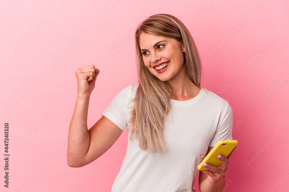Young russian woman holding a mobile phone isolated on pink background raising fist after a victory, winner concept. Young russian woman holding a mobile phone isolated on pink background raising fist after a victory, winner concept.
