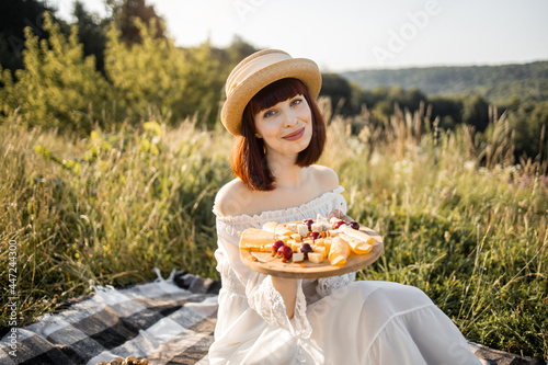 Portrait of cheerful happy young lady in dress and hat  sitting on the picnic blanket with round wooden plate with cheese and sweet cherries  enjoying vacation time