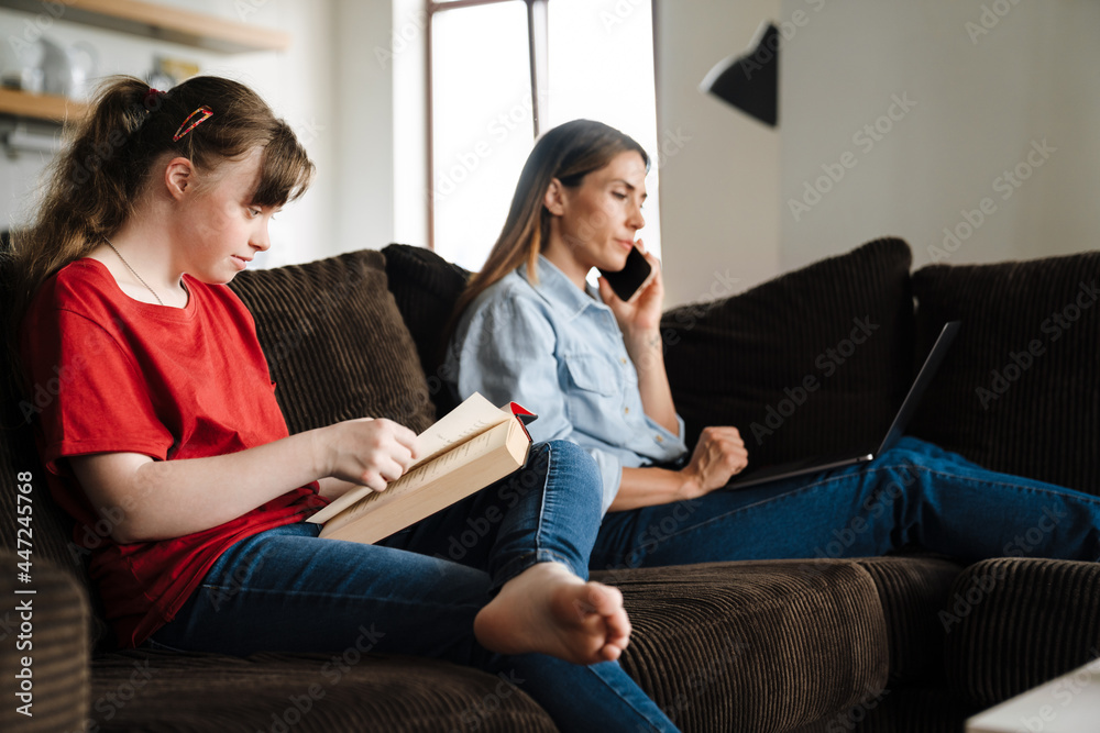 Girl with down syndrome reading book while sitting on sofa with her mother Girl with down syndrome reading book while sitting on sofa with her mother