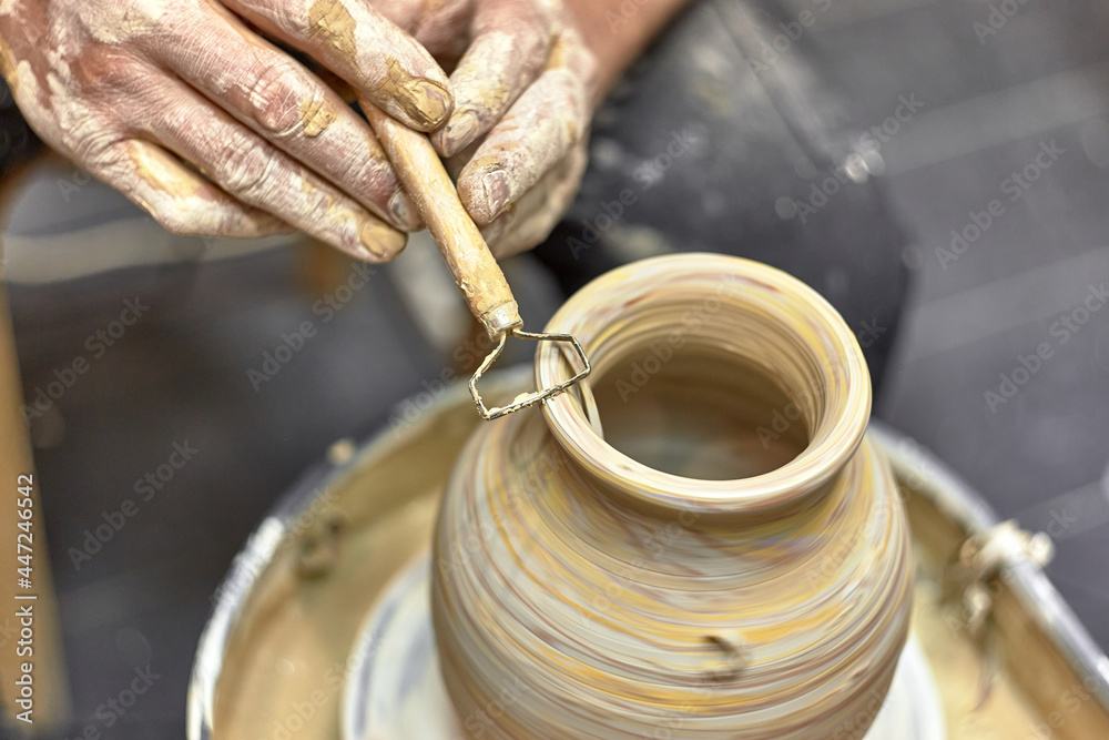 Hands of a ceramist in the process of leveling a finished vase of light clay on a potter's wheel using a specialized tool Hands of a ceramist in the process of leveling a finished vase of light clay on a potter's wheel using a specialized tool