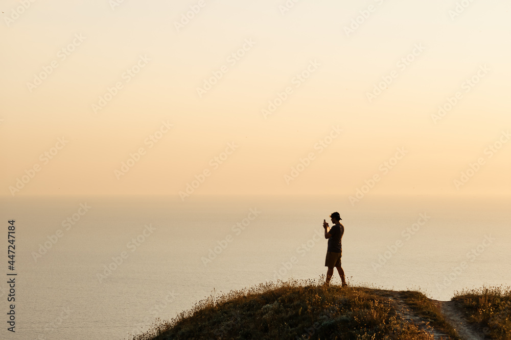 Silhouette of a young man taking pictures of the sea on a smartphone during sunset. Evening, summer travel on vacation