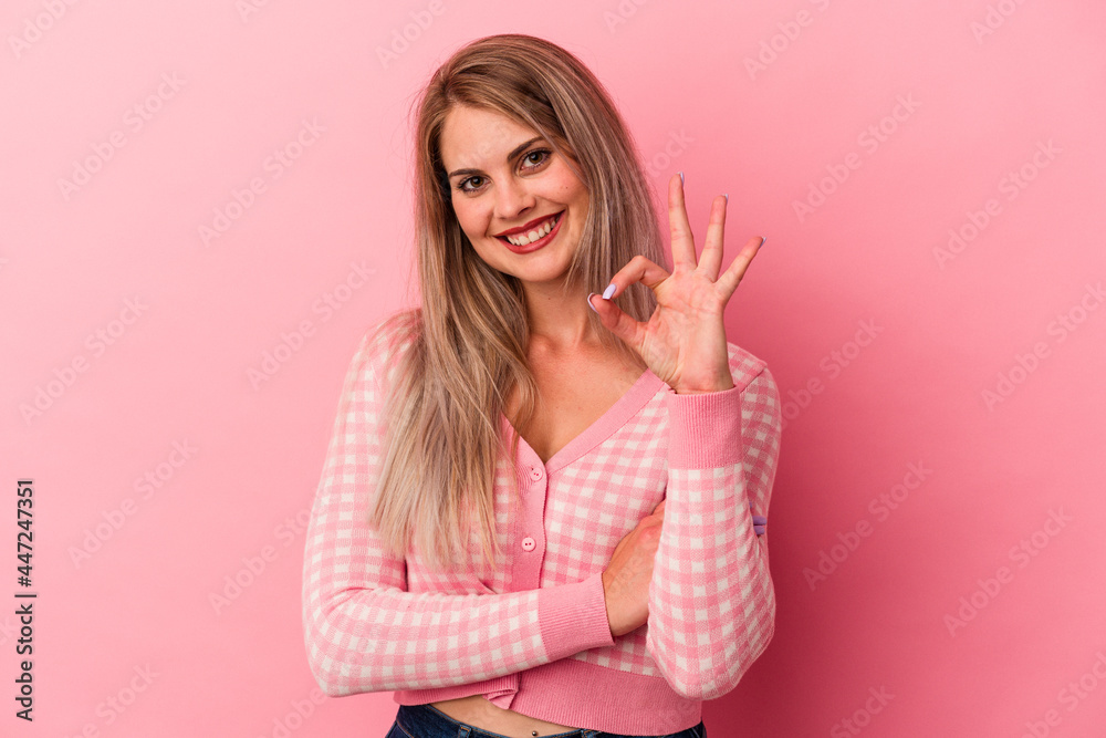 Young russian woman isolated on pink background winks an eye and holds an okay gesture with hand.