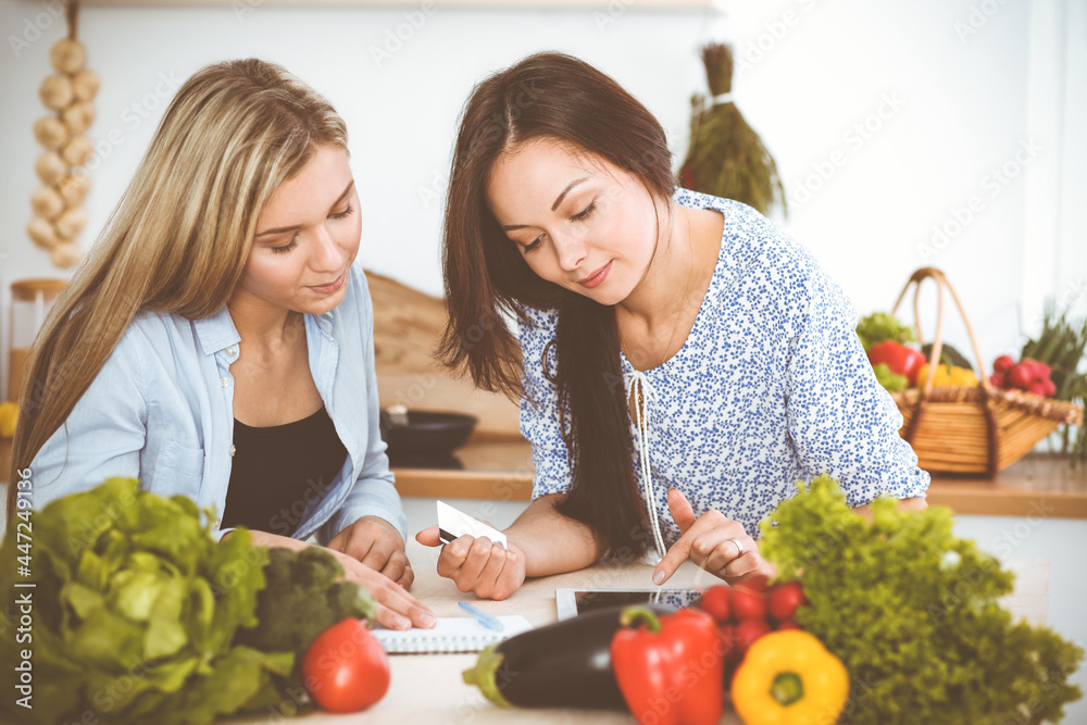 Two women friends choosing the recipe for a delicious meal while sitting at the table in the kitchen. Tablet pc is the best cookbook