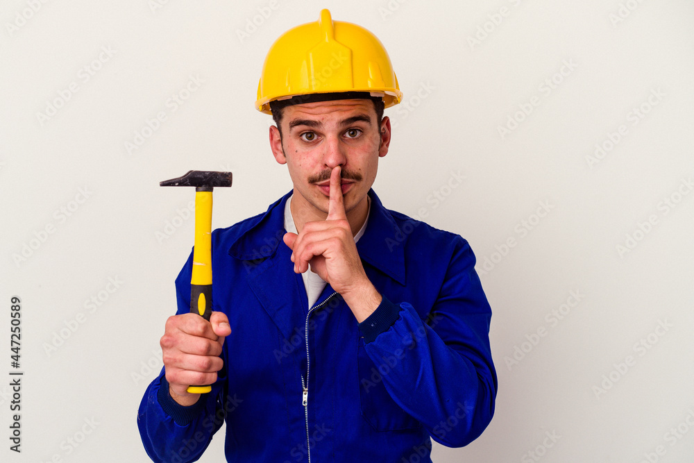 Young caucasian worker man holding a hammer isolated on white background keeping a secret or asking for silence. Young caucasian worker man holding a hammer isolated on white background keeping a secret or asking for silence.