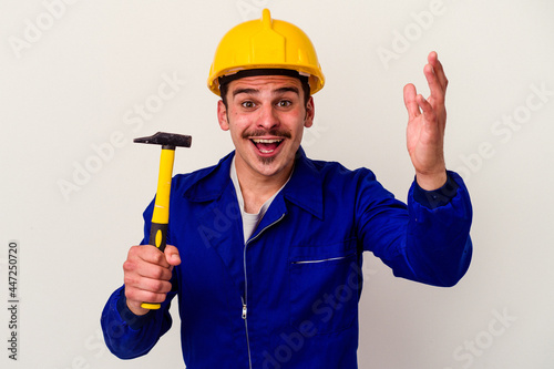 Young caucasian worker man holding a hammer isolated on white background receiving a pleasant surprise, excited and raising hands.