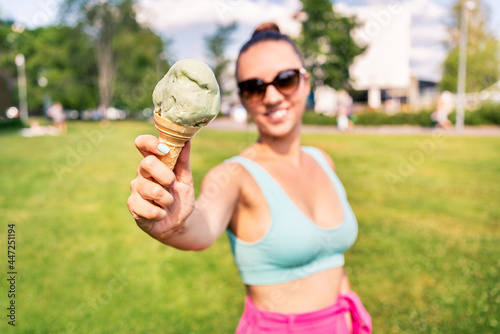 Fit slim woman showing ice cream cone in city park in summer. Happy smiling portrait of young laughing model and melting icecream. Stylish trendy bikini and sunglasses. Scoop of gelato. Outdoor joy. © terovesalainen