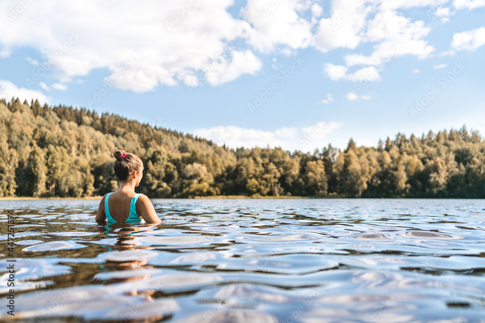 Fototapeta premium Lake in Finland. Woman swimming. Finnish nature, water and serene sky in summer. Outdoor bathing in Scandinavia after sauna. Green forest and beach in the morning or day. Lakeside back view portrait