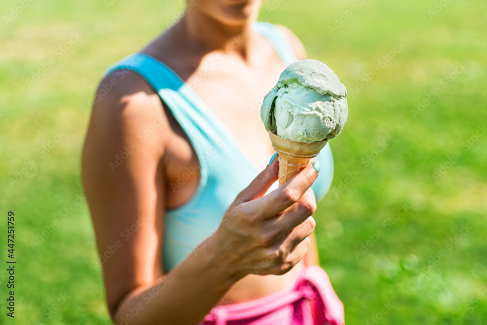 Ice cream cone in hand of a fit woman in summer. Gelato and colorful fashion in park. Retro vibes in outfit and icecream on sunny weekend. Eating and enjoying summertime vacation outside. Green grass. Ice cream cone in hand of a fit woman in summer. Gelato and colorful fashion in park. Retro vibes in outfit and icecream on sunny weekend. Eating and enjoying summertime vacation outside. Green grass.