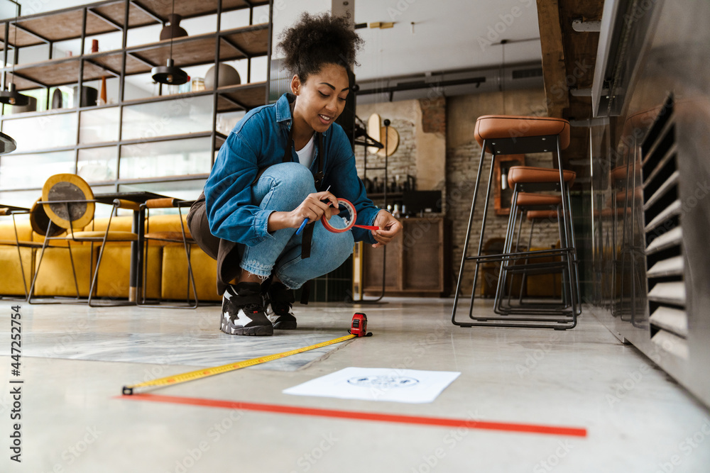 Obraz premium Black waitress in apron using tape measure while working at cafe