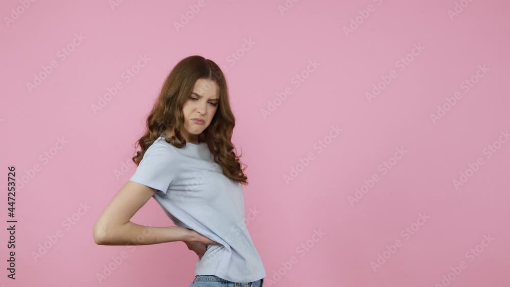 Backache. Tired overworked young woman in Gray t-shirt touching injured sore back, suffering spasms from torn muscles, pinched nerve, frowning face. Indoor studio shot isolated over pink background. Backache. Tired overworked young woman in Gray t-shirt touching injured sore back, suffering spasms from torn muscles, pinched nerve, frowning face. Indoor studio shot isolated over pink background.