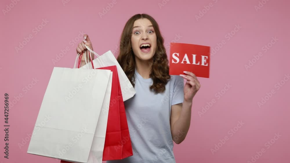 Trendy teenager girl raising shopping bags and card with sale inscription, looking satisfied with purchase, enjoying seasonal discounts. Indoor studio shot isolated over pink background. Trendy teenager girl raising shopping bags and card with sale inscription, looking satisfied with purchase, enjoying seasonal discounts. Indoor studio shot isolated over pink background.