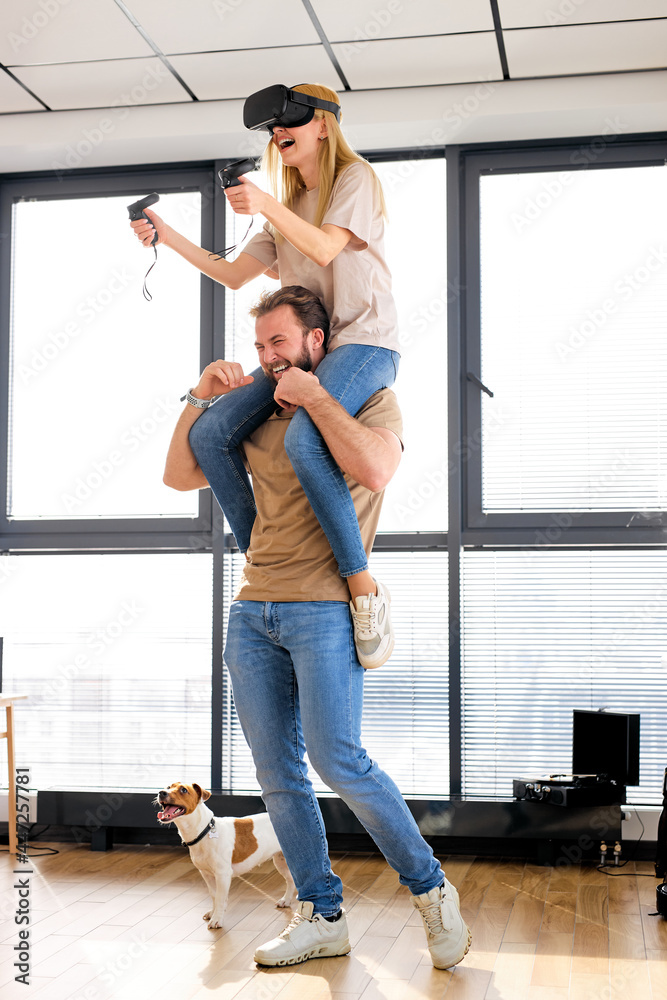 Fototapeta premium Couple caucasian man and woman play the game console with VR glasses at home, man holding wife on shoulders. Excited people having fun with new trends technology. Gaming concept. Video game
