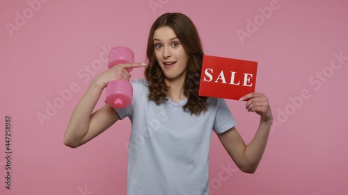 Beautiful teenager girl wearing casual style attire lifting up dumbbell and showing red card with sale inscription, offers discounts for sport goods. Indoor studio shot isolated over pink background.