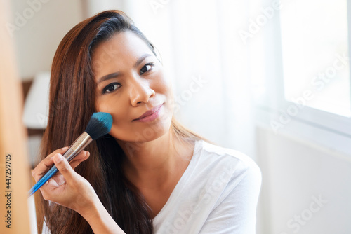 Smiling lovely young woman applying powder blush on her cheeks when getting ready in the morning