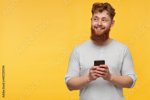 young redhead male with a big beard smiles and holds smartphone in his hands. looking aside to the right at copy space. isolated over yellow background © timtimphoto