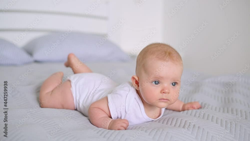 Indoor shot of charming newborn girl wearing white bodysuit having tummy time, lying on bed on gray blanker, studying world around, infant developing, childcare. Indoor shot of charming newborn girl wearing white bodysuit having tummy time, lying on bed on gray blanker, studying world around, infant developing, childcare.