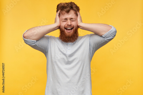 young redhead bearded man with, keeps his eyes closed, holding head with both hands, screaming while feels pain. isolated over yellow background.