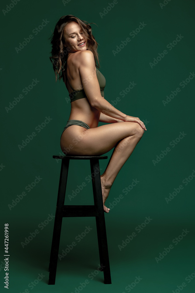 Fototapeta premium One young attractive girl, female model in green lingerie, swimming suit sitting high chair isolated over dark green studio background. Natural beauty