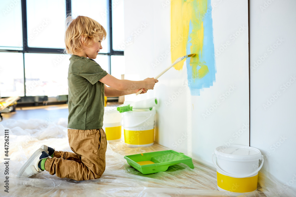 Funny little caucasian kid boy painting the wall with color in new house.  Family repair apartment home. Happy child paints the wall, applying with  paintbrush, having fun with brush, indoors. Stock Photo |
