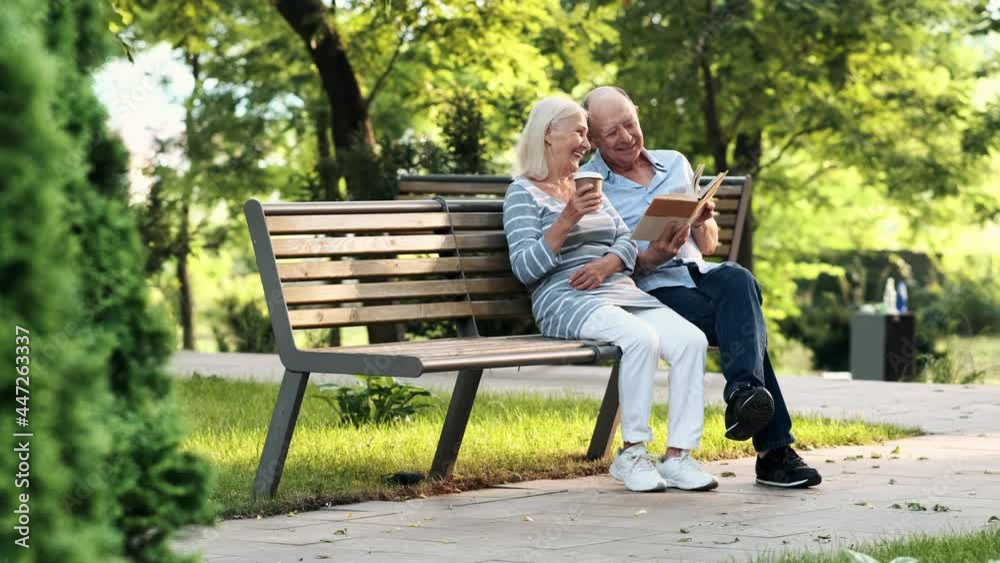 A happy elderly couple man and woman are sitting on the bench while reading book in the park