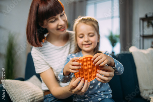 Stress relief squeeze sensory pop it and simple dimple toys for kids. Lovely pretty mother sitting on sofa at home and playing pop pit game with her cheerful little daughter