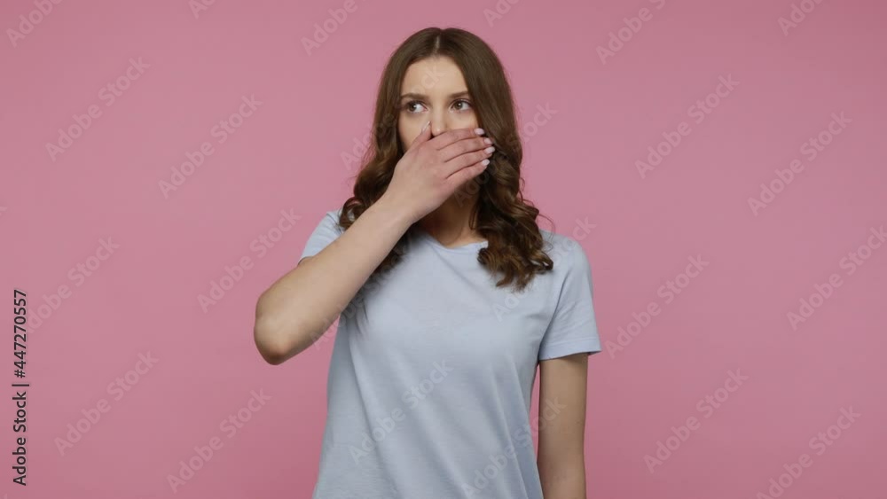 Terrified wavy haired teenager girl covering her mouth with palm to keep silence, surprised by horrible truth, promising don't tell secret to anyone. Indoor studio shot isolated over pink background. Terrified wavy haired teenager girl covering her mouth with palm to keep silence, surprised by horrible truth, promising don't tell secret to anyone. Indoor studio shot isolated over pink background.