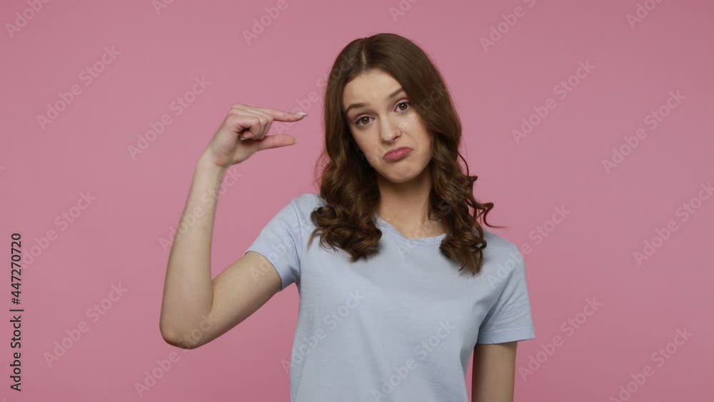 Pretty teenager girl in casual T- shirt showing small size, looking with disappointed expression, meaning little size, low rating, measuring scale. Indoor studio shot isolated over pink background. Pretty teenager girl in casual T- shirt showing small size, looking with disappointed expression, meaning little size, low rating, measuring scale. Indoor studio shot isolated over pink background.