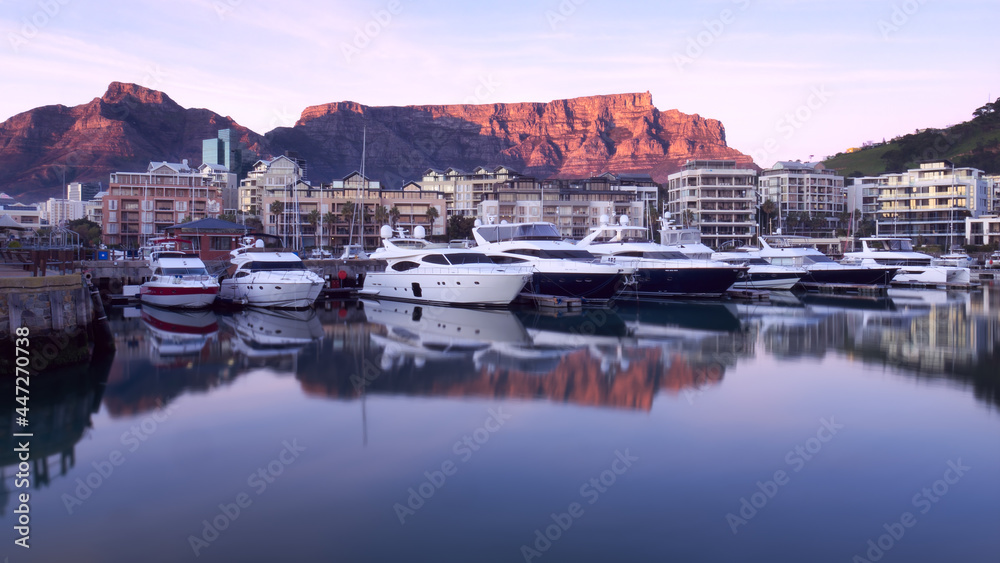 Fototapeta premium Table Mountain sunrise is reflected in the still waters of a marina for luxury motor yachts in Cape Town, South Africa