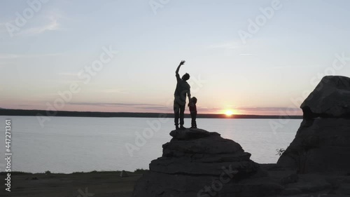 The drone captures a man and his son, who are standing on the top of a cliff and looking at the view. The camera gradually goes to the side. You can see the pink sky, the sun is setting and the lake photo