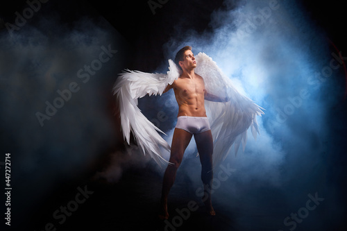 Handsome young athletic man with a bare torso who looks like an angel with white wings. Model dancer posing in a dark studio on black background