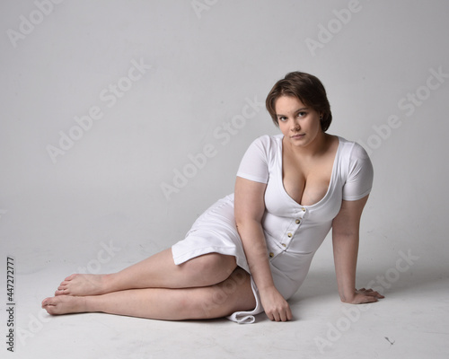 Full length portrait of young plus sized woman with short brunette hair, wearing l tight white body con dress, kneeling pose with gestural hands on ground with light studio background.