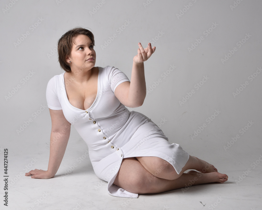 Full length portrait of young plus sized woman with short brunette hair,  wearing l tight white body con dress, kneeling pose with gestural hands on  ground with light studio background. Stock Photo