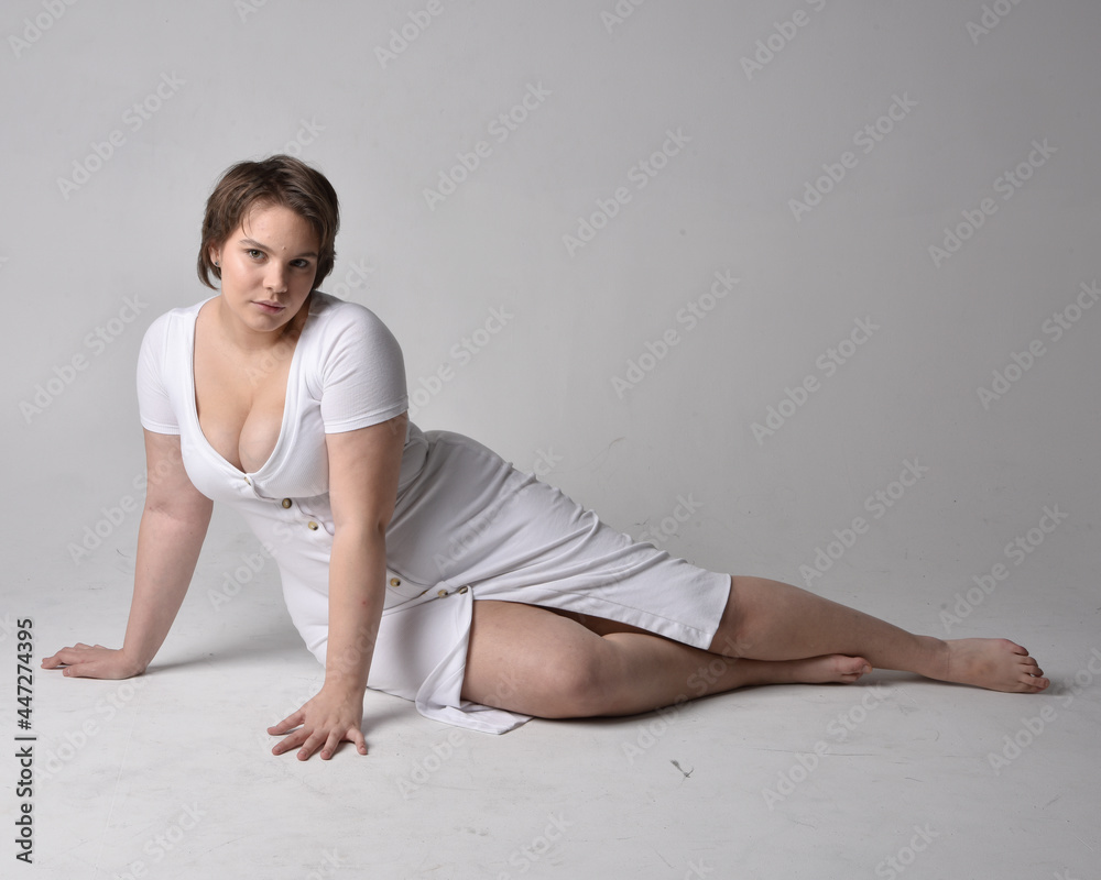 Full length portrait of young plus sized woman with short brunette hair, wearing l tight white body con dress, kneeling pose with gestural hands on ground with light studio background. Full length portrait of young plus sized woman with short brunette hair, wearing l tight white body con dress, kneeling pose with gestural hands on ground with light studio background.