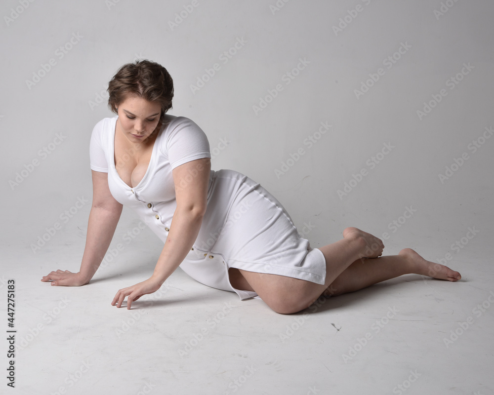 Fototapeta premium Full length portrait of young plus sized woman with short brunette hair, wearing l tight white body con dress, kneeling pose with gestural hands on ground with light studio background.